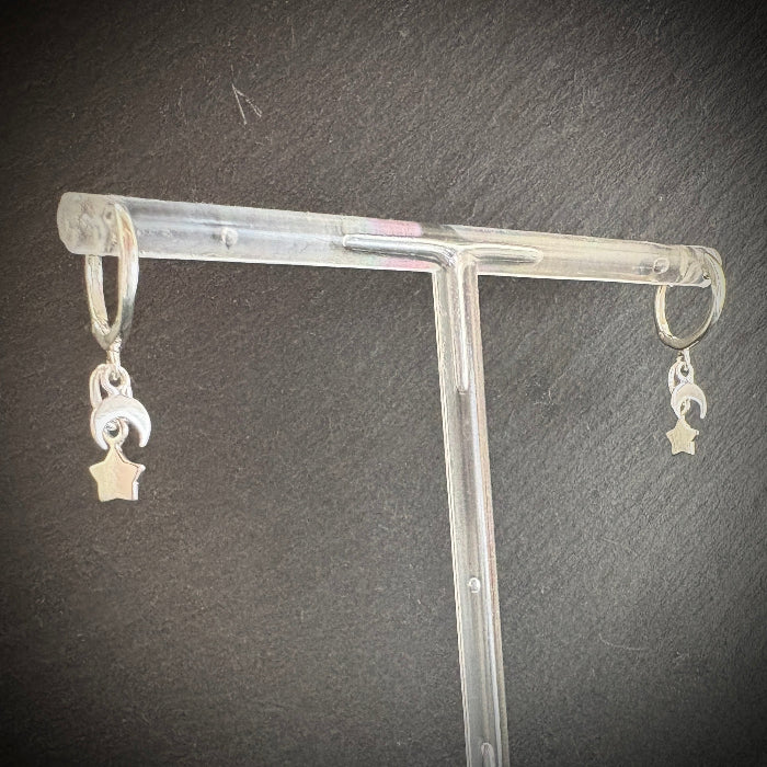 Silver earrings with star and moon designs on a clear stand against a dark background