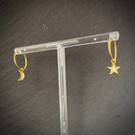Gold hoop earrings with star and moon designs on a clear stand against a dark background