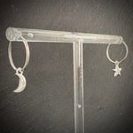 Silver hoop earrings with moon and star charms on a clear stand against a dark background